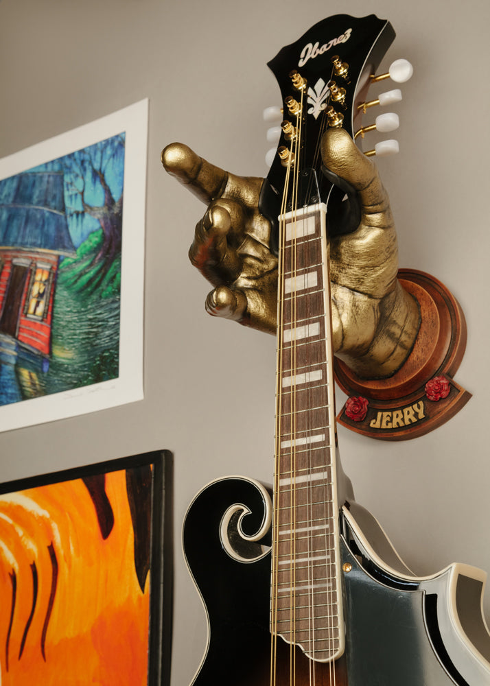 Jerry Garcia gold hand guitar hanger with missing middle finger holding black mandolin. Art work next to the hanging guitar. 